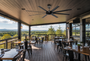 A large covered outdoor dining patio with wooden tables and chairs, a ceiling fan, and panoramic windows overlooking a forested mountain landscape.
